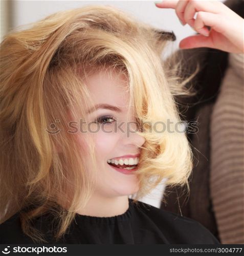 Beautiful smiling girl with blond wavy hair by hairdresser. Hairstylist combing female client. Young woman in hairdressing beauty salon. Hairstyle.
