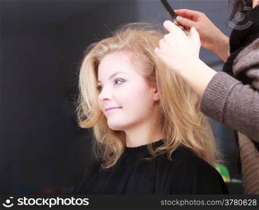 Beautiful smiling girl with blond wavy hair by hairdresser. Hairstylist combing female client. Young woman in hairdressing beauty salon. Hairstyle.