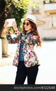 Beautiful smiling hipster woman wear vintage sunglasses, jeans shirt, white hat and jacket take a picture of herself with digital tablet. Selfie style. Toned in warm colors. Outdoors shot, lifestyle
