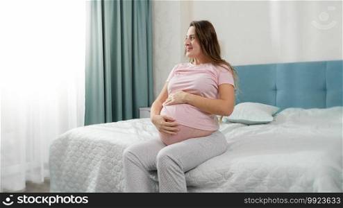 Beautiful smiling pregnant woman in pajamas sitting on bed and stroking big belly at morning.. Beautiful smiling pregnant woman in pajamas sitting on bed and stroking big belly at morning