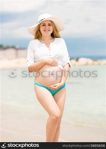 Beautiful smiling pregnant woman in white shirt posing on beach at sunny day