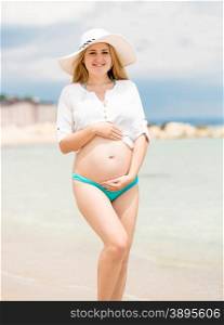 Beautiful smiling pregnant woman in white shirt posing on beach at sunny day