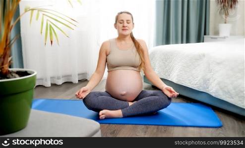 Beautiful smiling pregnant woman practicing yoga sitting on fitness mad in lotus pose and breathing deeply. Concept of healthcare and sports during pregnancy.. Beautiful smiling pregnant woman practicing yoga sitting on fitness mad in lotus pose and breathing deeply. Concept of healthcare and sports during pregnancy