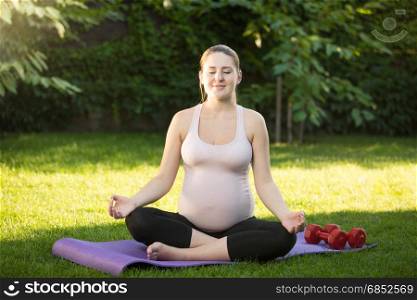 Beautiful smiling pregnant woman sitting in lotus pose on grass at park