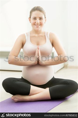 Beautiful smiling pregnant woman sitting in yoga lotus position on fitness mat