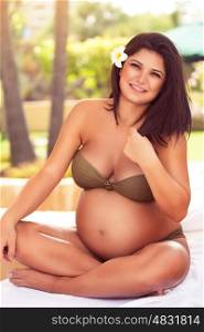 Beautiful smiling pregnant woman sitting on the beach, enjoying hot summer on the tropical resort, happy healthy pregnancy