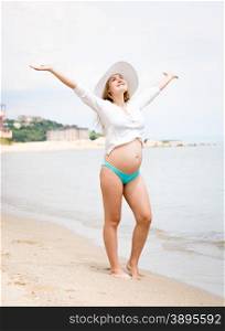 Beautiful smiling pregnant woman standing on beach and holding hands up