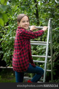 Beautiful smiling teenage girl in red checkered shirt climbing up stepladder at garden