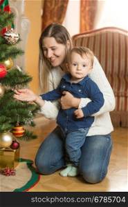 Beautiful smiling woman decorating Christmas tree with her 10 months old baby son