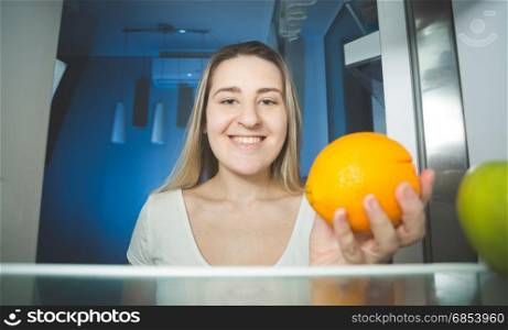 Beautiful smiling woman holding orange and looking inside the refrigerator
