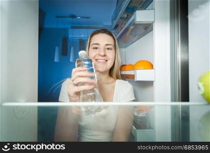Beautiful smiling woman taking water from fridge and drinking it. View from inside of open refrigerator