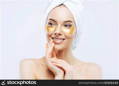 Beautiful smiling young woman applies hydrogel moisturizing pads under eyes, smiles tenderly and looks aside, wears wrapped towel on head, enjoys hygiene procedures, stands shirtless indoor.