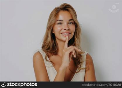 Beautiful smiling young woman keeps index finger over lips tells secret spreads rumors has makeup clean healthy skin looks directly at camera poses against grey background. Hush be quiet please. Beautiful smiling young woman keeps index finger over lips tells secret spreads rumors