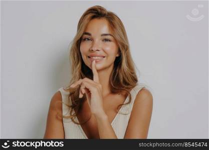Beautiful smiling young woman keeps index finger over lips tells secret spreads rumors has makeup clean healthy skin looks directly at camera poses against grey background. Hush be quiet please. Beautiful smiling young woman keeps index finger over lips tells secret spreads rumors
