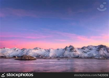 Beautiful snow covered mountains and colorful sky reflected in water at night. Winter landscape with sea, snowy rocks, sky, pink clouds, reflection at sunset. Lofoten islands, Norway at dusk. Nature