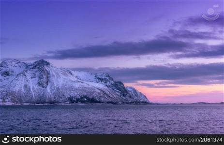Beautiful snow covered mountains and violet sky reflected in water at dusk. Winter landscape with sea, snowy rocks, purple sky, reflection, at sunset. Lofoten islands, Norway at twilight. Nature