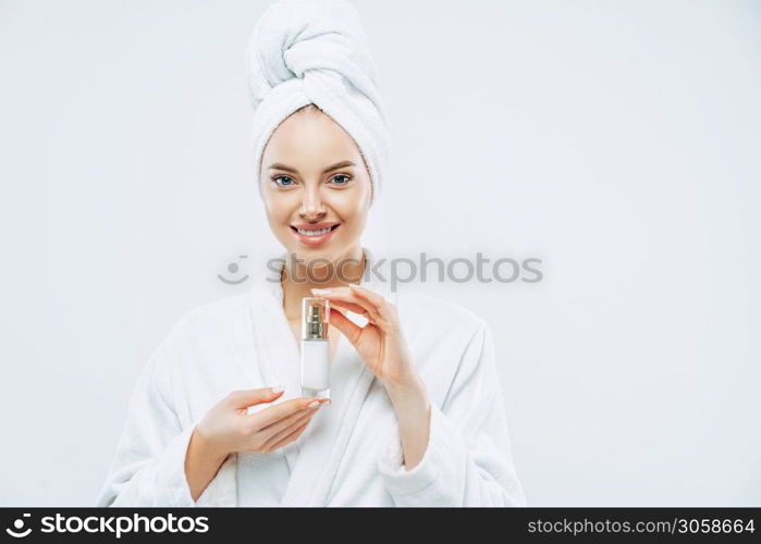 Beautiful spa young woman with healthy fresh skin applies anti aging lotion or cosmetic cream, uses day moisturizer, stands indoor, dressed in bath robe and towel, takes shower before going out