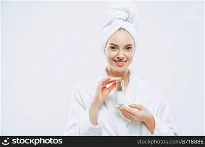 Beautiful spa young woman with healthy fresh skin applies anti aging lotion or cosmetic cream, uses day moisturizer, stands indoor, dressed in bath robe and towel, takes shower before going out
