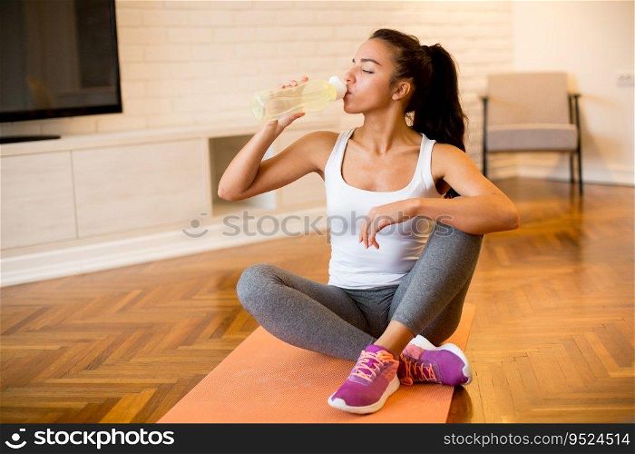 Beautiful sports woman drinking water while resting and sitting on yoga mat at home