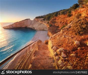 Beautiful stairs on sandy beach at sunset. Porto Katsiki, Lefkada island, Greece. Colorful landscape with blue sea, beautiful rocks in water, steps, empty beach, stones, green trees, sky, sunlight