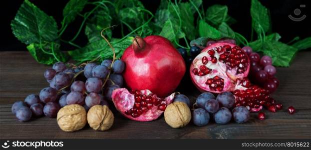 Beautiful still life with red pomegranates, blue ripe grapes and walnuts on a dark wooden surface, horizontal image