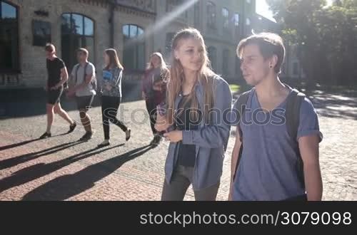 Beautiful student couple walking on univesity campus, holding books and chatting. Joyful hipster college friends going to lesson through park outside university building and talking with group of classmates on background. Slow motion. Stabilized shot