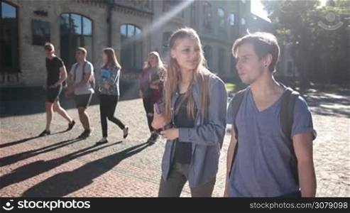 Beautiful student couple walking on univesity campus, holding books and chatting. Joyful hipster college friends going to lesson through park outside university building and talking with group of classmates on background. Slow motion. Stabilized shot