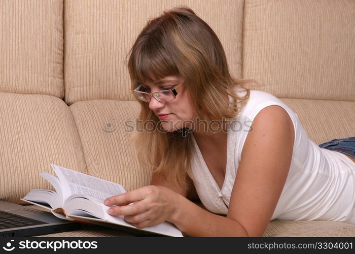 Beautiful student girl lying on the sofa with book and laptop.