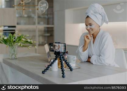 Beautiful successful female blogger records tutorial video talks with followers looks at camera of smartphone on tripod applies beauty patches under eyes dicuss how to care about skin and complexion