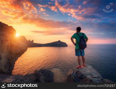 Beautiful summer landscape with standing man with backpack on the stone at the ocean against the colorful sky with clouds at sunset. Travel background. Sport, lifestyle. Tourism