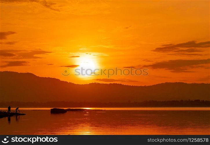 Beautiful sunrise sky above the mountain at reservoir. People are fishing with a fishing rod on the river. Landscape of reservoir and mountain with orange sunrise sky. Silhouette life in the morning.