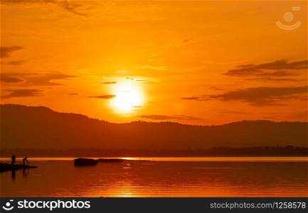 Beautiful sunrise sky above the mountain at reservoir. People are fishing with a fishing rod on the river. Landscape of reservoir and mountain with orange sunrise sky. Silhouette life in the morning.