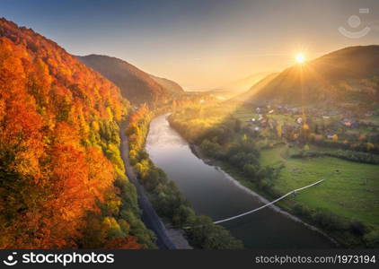 Beautiful sunset in mountains in autumn. Aerial view of forest with red trees on mountain, river, road, bridge, green meadow, village in fall in Ukraine. Colorful rural landscape. Top view. Nature