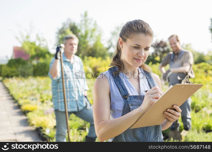 Beautiful supervisor writing on clipboard with gardeners standing in background at plant nursery