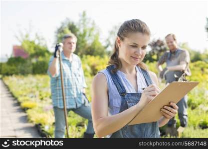 Beautiful supervisor writing on clipboard with gardeners standing in background at plant nursery