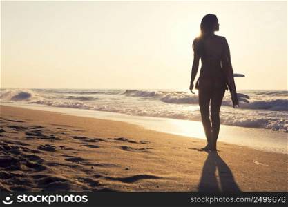 Beautiful surfer girl walking in the beach with her surfboard at sunset
