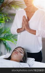 Beautiful teenage girl lying with her eyes closed and having Reiki healing treatment. Reiki Practitioner standing and praying above patient’s body. 