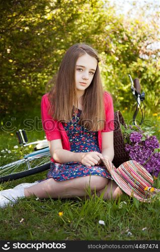 Beautiful teenage girl sitting in a field with a bicycle with a basket of flowers in background looking straight to camera