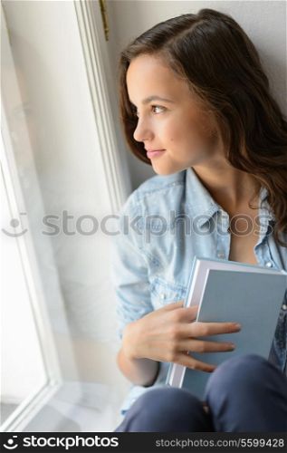 Beautiful teenage girl student holding book looking out the window