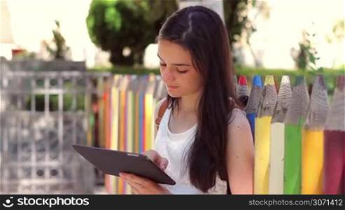 Beautiful teenage girl student using tablet pc in front of the school showing it at camera.