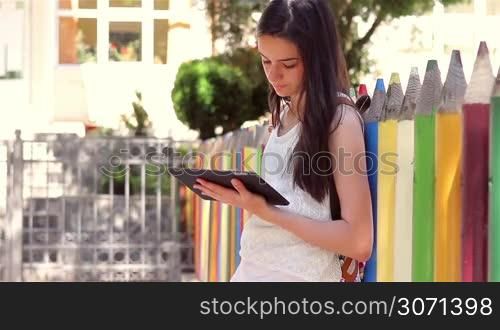 Beautiful teenage girl student writing or reading something from tablet pc in front of the school.