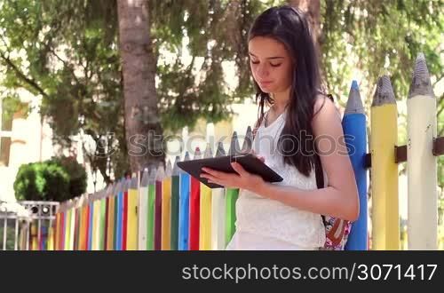 Beautiful teenage girl student writing or reading something from tablet pc in front of the school.
