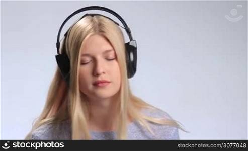 Beautiful teenage girl with closed eyes swaying dreamily to her music streaming with headphones on white background. Young woman opening her eyes and smiling sheepishly, touching earcups then continues listening to music with closed eyes.
