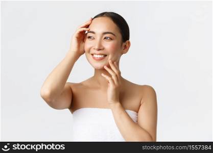 Beautiful tender asian girl in towel looking away pleased, touching clean face, no acne, enjoying day in spa, standing bathroom over white background, apply cleanser or skincare product.. Beautiful tender asian girl in towel looking away pleased, touching clean face, no acne, enjoying day in spa, standing bathroom over white background, apply cleanser or skincare product