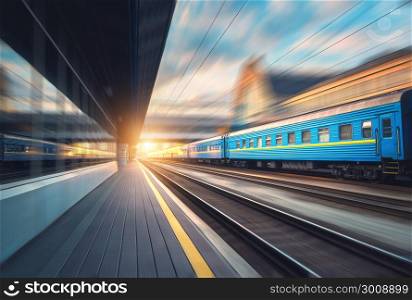 Beautiful train with blue wagons in motion at the railway station at sunset. Industrial view with modern train, railroad, railway platform, buildings, cloudy sky with motion blur effect. Concept