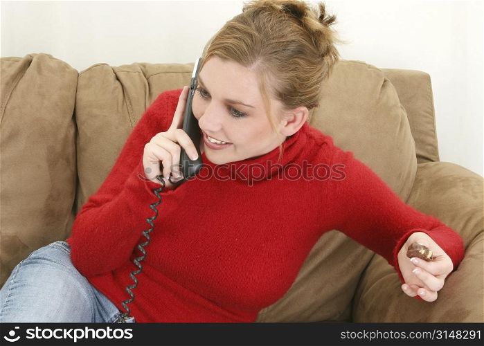 Beautiful twenty-something woman in red sweater sitting on couch eating chocolate and talking on phone.