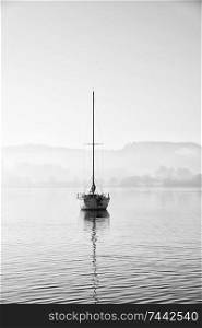 Beautiful unplugged landscape image of sailing yacht sitting still in calm lake water in Lake District during peaceful misty Autumn Fall sunrise
