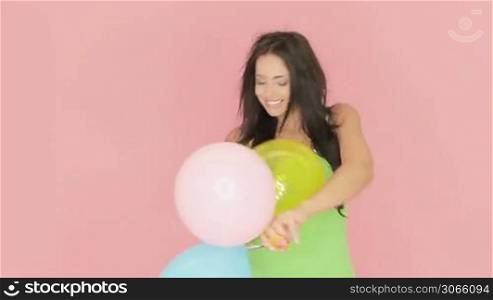 Beautiful vivacious woman playing with a colourful bunch of party balloons against a pink studio background