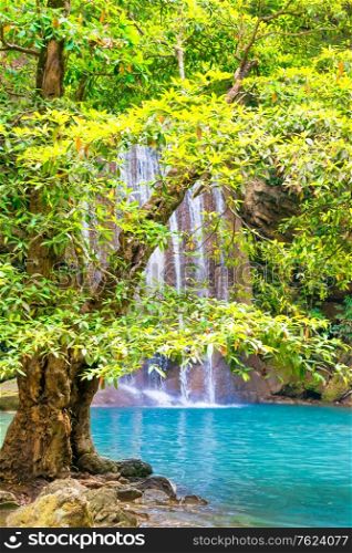 Beautiful waterfall in tropical jungle forest with big green tree on foreground and emerald lake. Nature landscape of Erawan National park, Kanchanaburi, Thailand