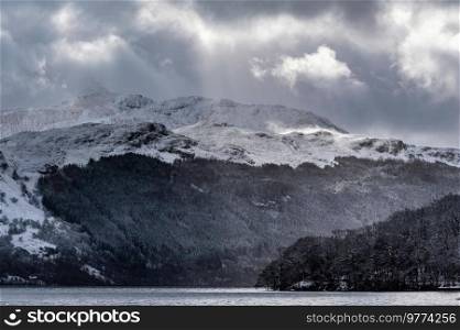 Beautiful Winter landscape image of snow covered trees on shores of Loch Lomond with Ben Lomond mountain looming in background